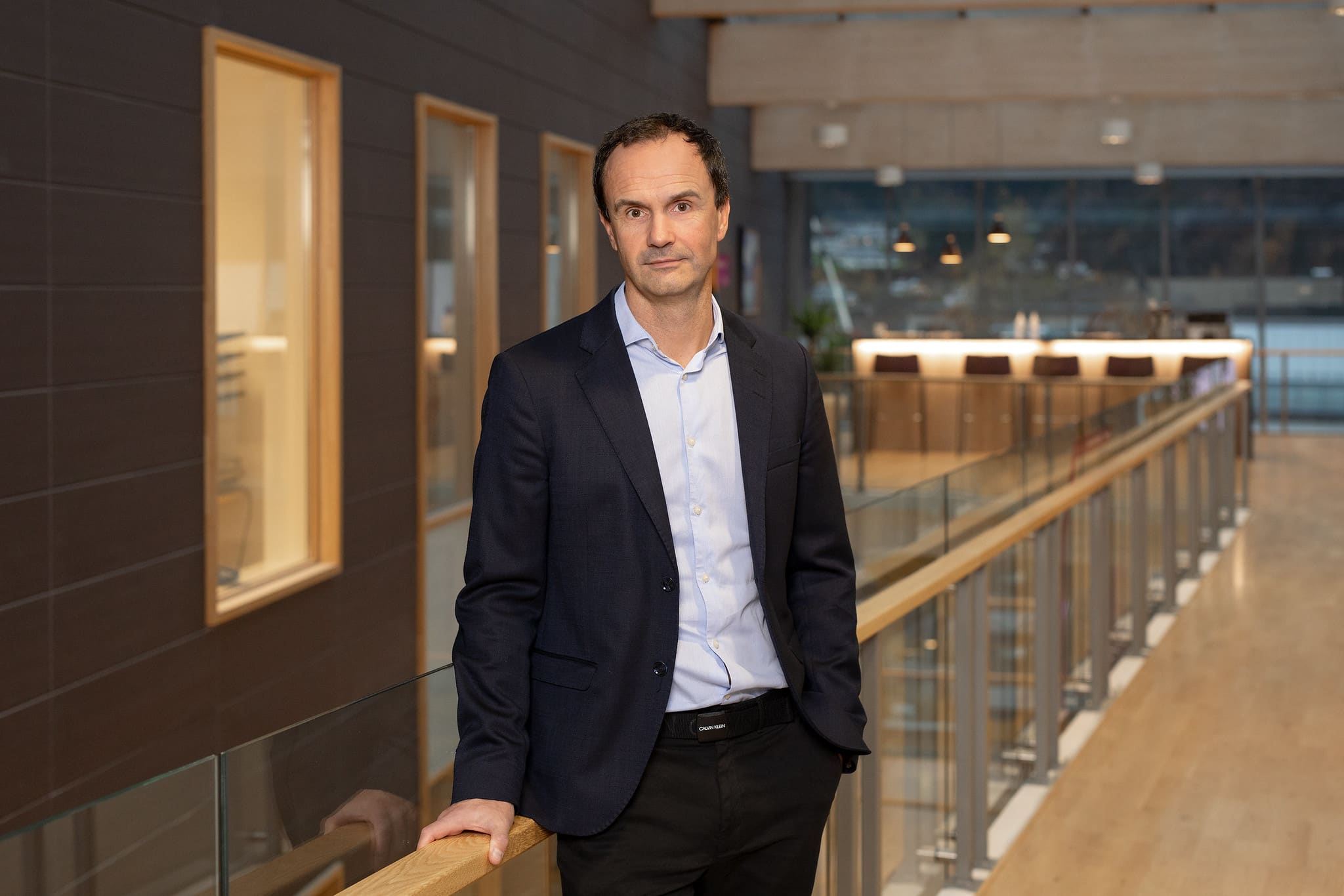 Bilde av Tore Bell, som er avdelingsdirektør i Lotteritilsynet. Han har på seg svart dress og en lås blå skjorte, og står ved et gelender inne i lokalene til Lotteri- og stiftelsestilsynet.